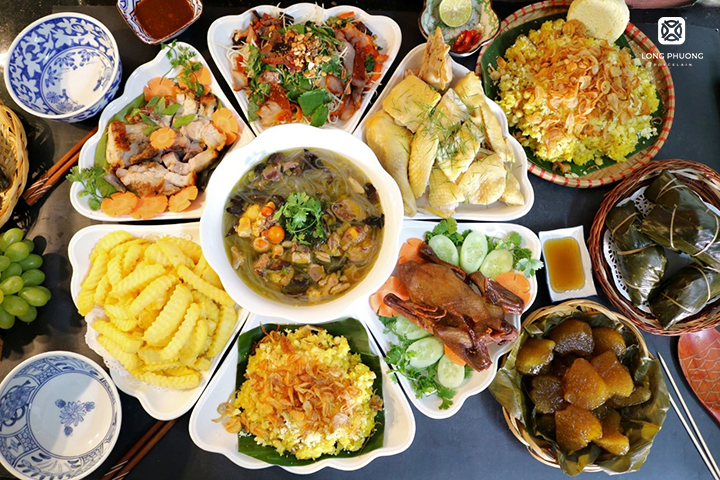 A tray of food prepared for a death anniversary — lavish and diverse — the family stands in prayer before eating