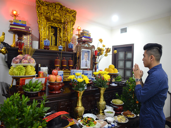 A young Vietnamese person lighting incense at an altar inside a modern apartment — the juxtaposition of a smartphone and traditional incense