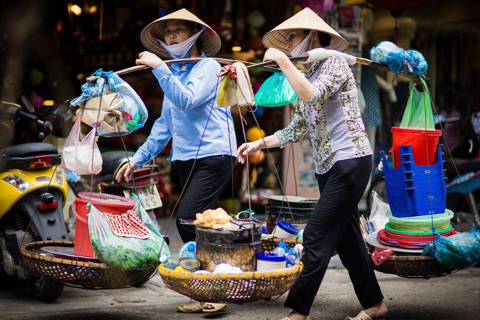 A Vietnamese woman carrying a yoke of goods on the street — a smile, a heavy burden, familiar footsteps