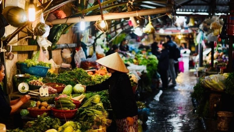 A bustling traditional Vietnamese market — vibrant colors of vegetables, buyers and sellers, chaotic noise