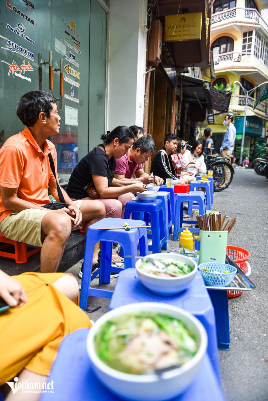 A sidewalk Pho stall in Hanoi in the early morning — low tables and chairs, steaming bowls, a blurred atmosphere