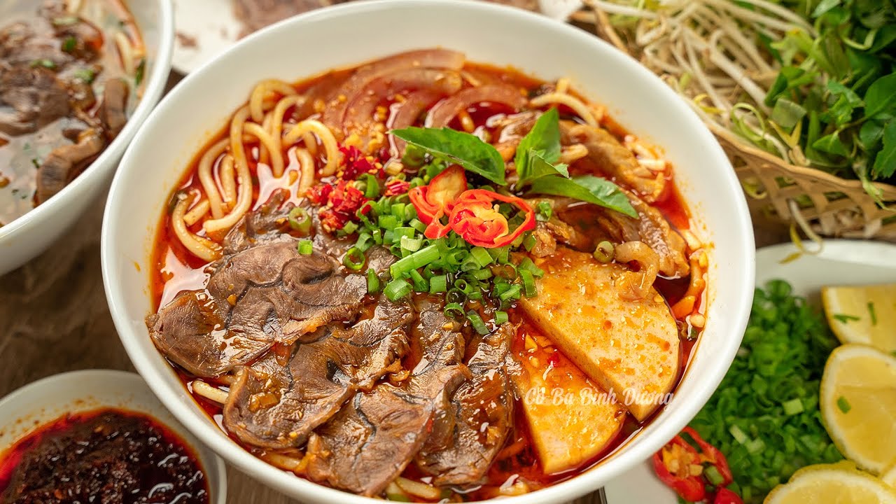 A blazing red bowl of Hue beef noodle soup — crimson broth, crab cake, lemongrass stalks